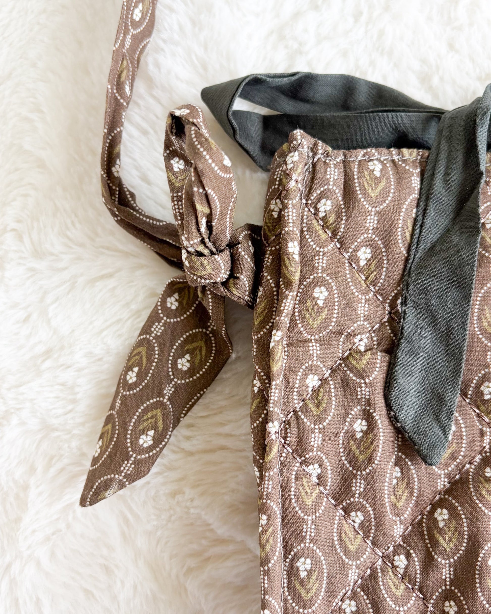 A close-up of a brown patterned fabric bag or satchel with a matching strap tied in a knot, resting on a white faux fur surface.