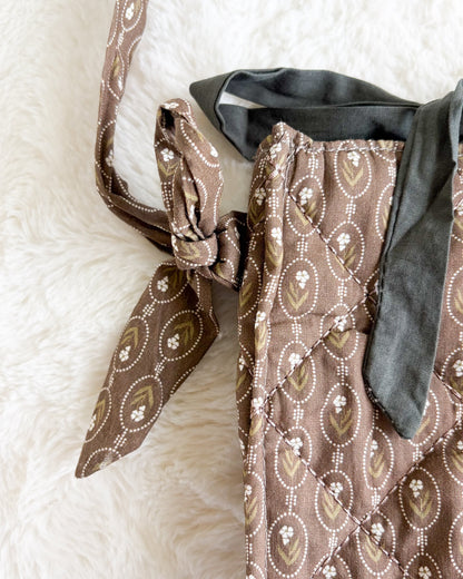 A close-up of a brown patterned fabric bag or satchel with a matching strap tied in a knot, resting on a white faux fur surface.