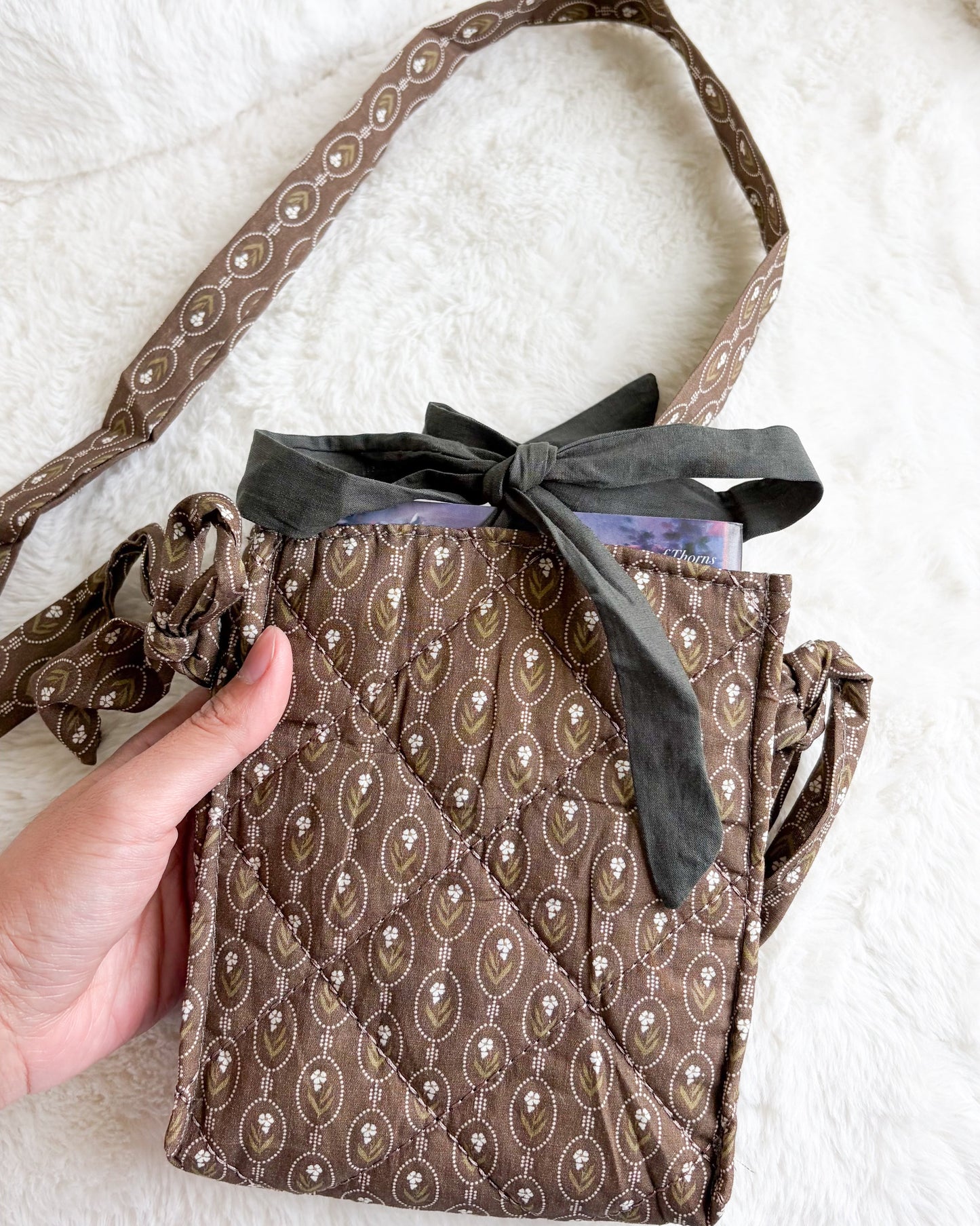 A brown quilted crossbody bag with a paisley pattern, featuring a dark gray ribbon bow at the top and a matching patterned strap, held by a hand against a white faux fur background.