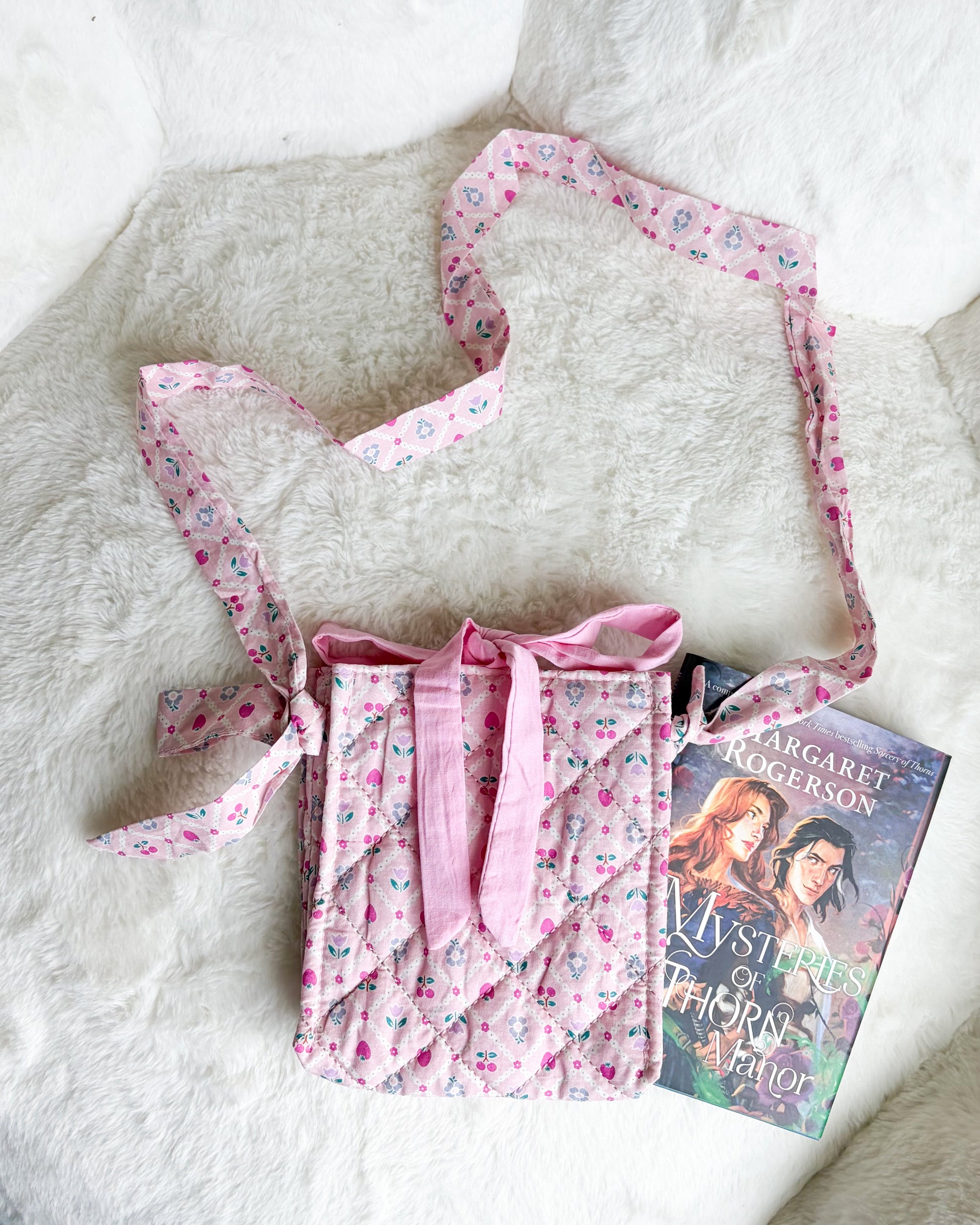 A pink quilted fabric crossbody bag with a floral print and matching strap, styled on a white faux fur surface next to a book.