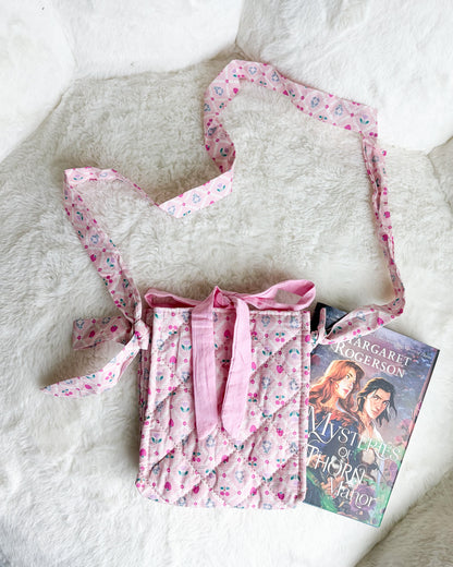 A pink quilted fabric crossbody bag with a floral print and matching strap, styled on a white faux fur surface next to a book.