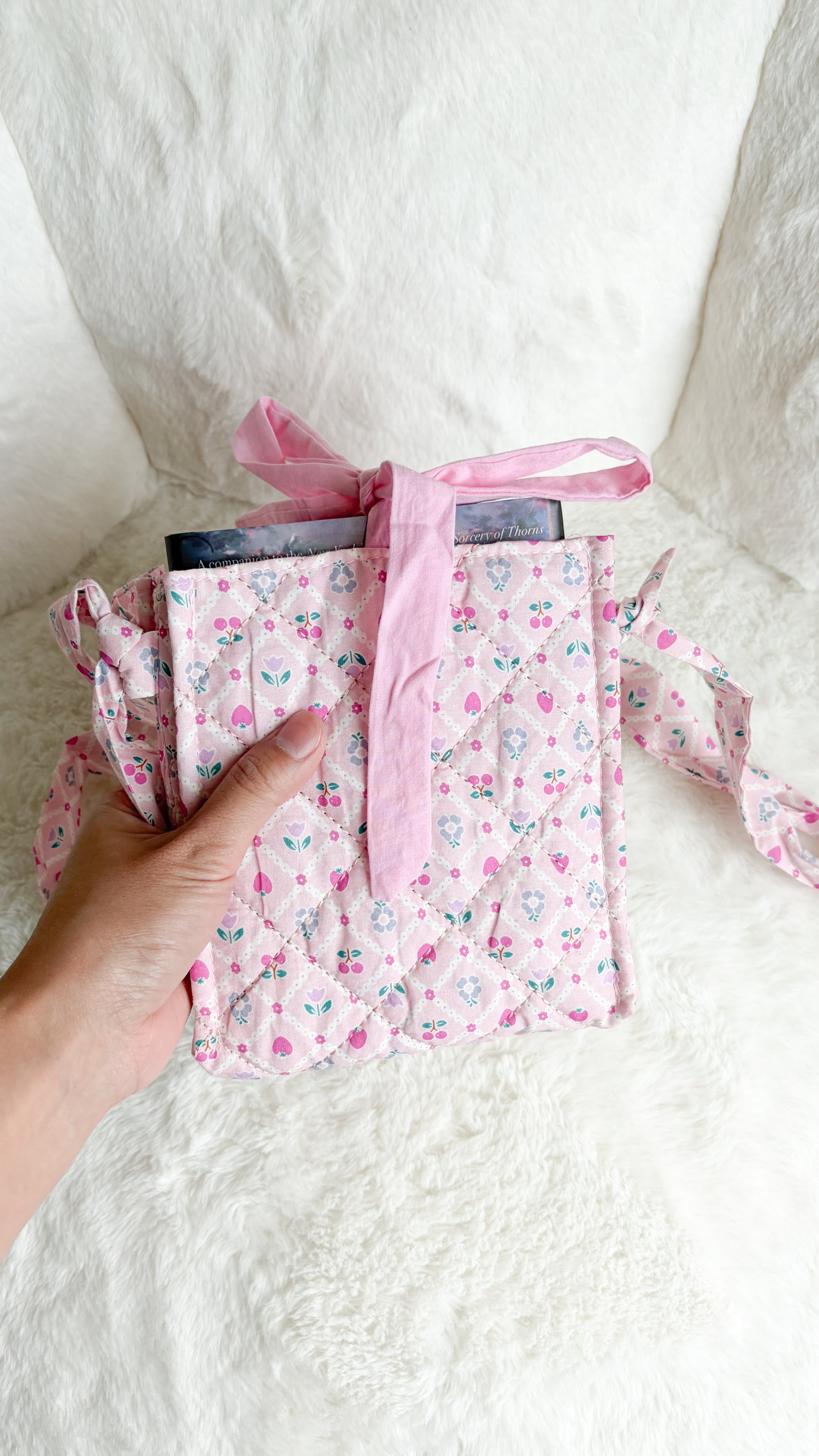 A hand holding a small pink floral fabric pouch or organizer with a matching pink ribbon bow, set against a soft white textured background.