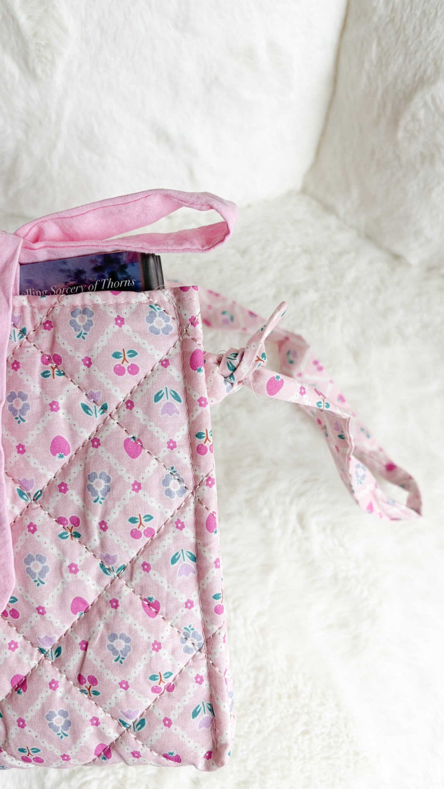 A pink quilted fabric tote bag with a cherry and floral pattern, featuring a pink ribbon handle, resting on a white textured blanket.