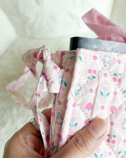 A close-up of a hand holding a light pink quilted fabric bag or pouch with a floral print of strawberries, hearts, and leaves, featuring a matching fabric tie closure.