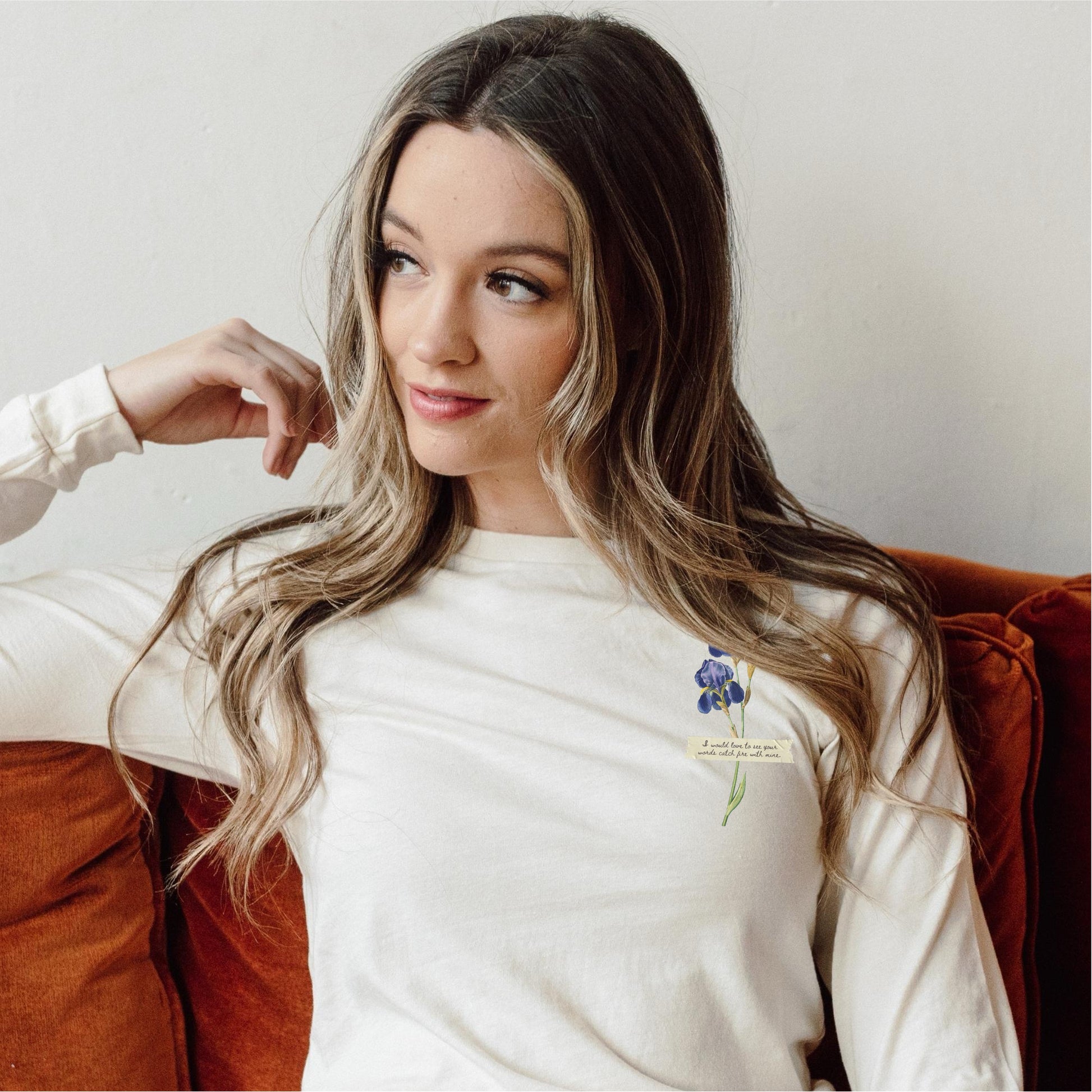 Woman wearing cream long sleeve tee with purple flower and bookish quote print, sitting on couch