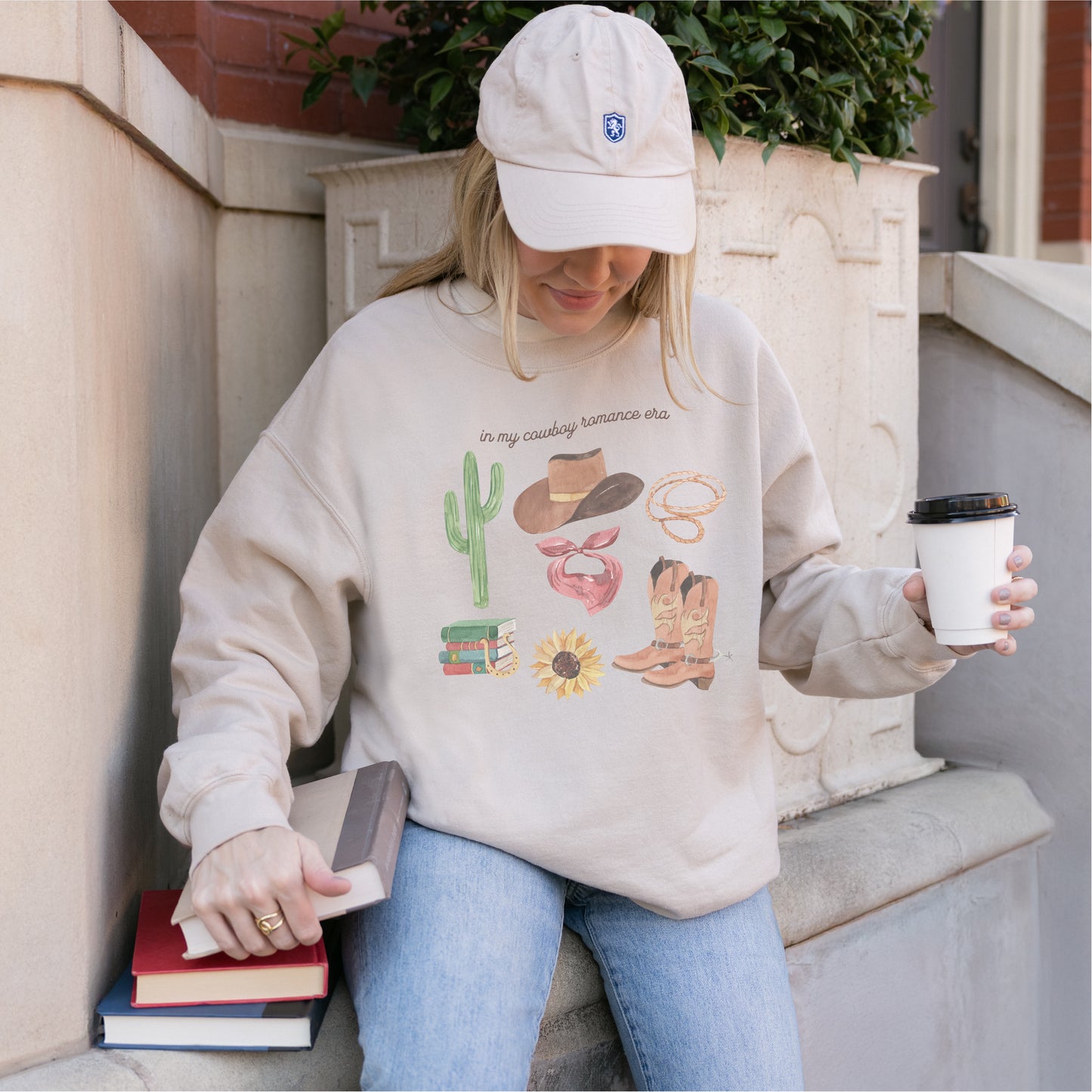 Woman in beige 'In My Cowboy Romance Era' sweatshirt with western and bookish graphics, holding coffee and books.