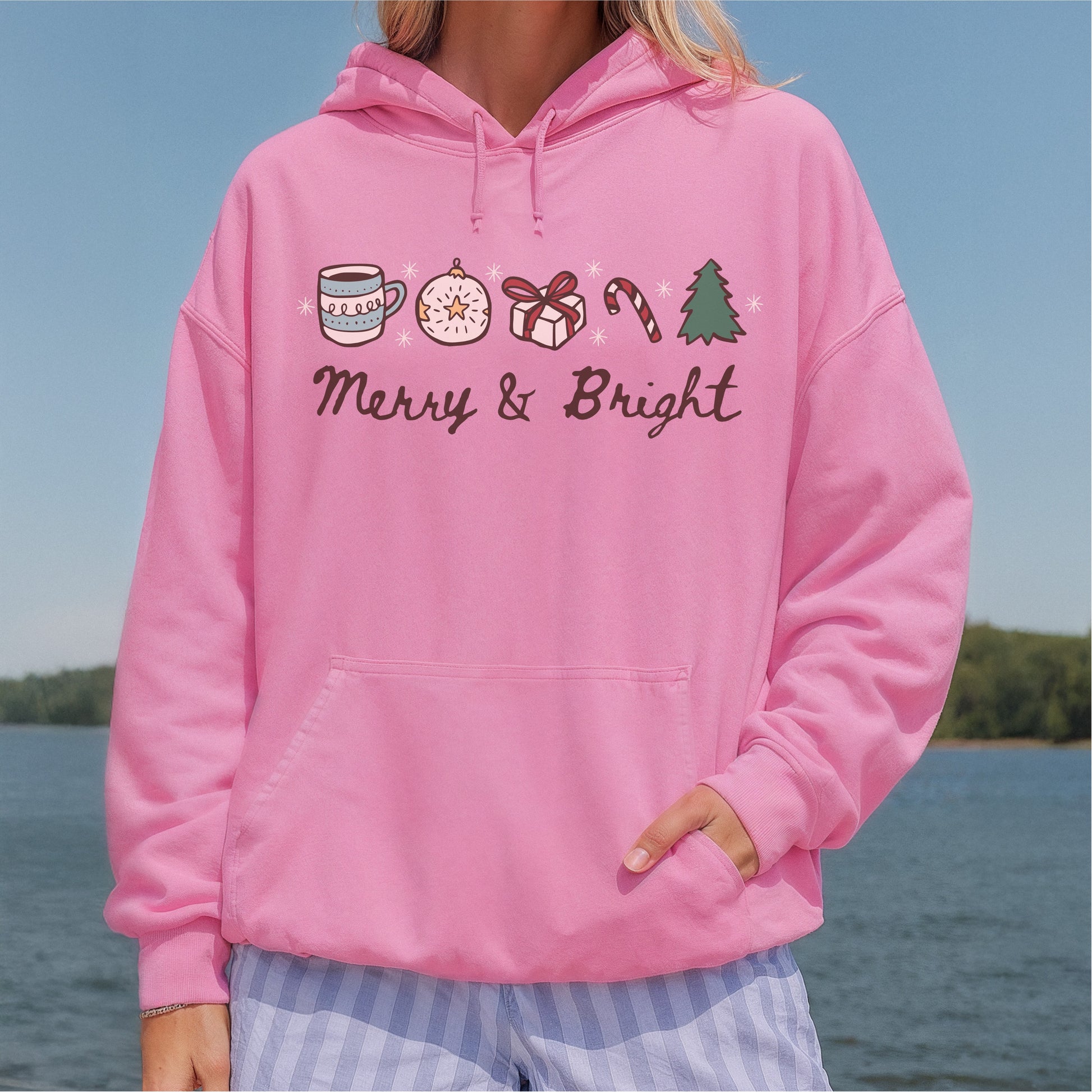 Pink hoodie with Merry & Bright holiday design, featuring festive icons, worn outdoors.