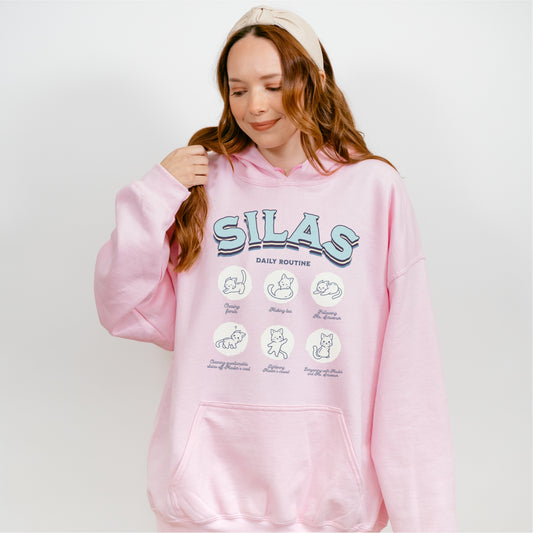 Woman wearing a pink Silas Daily Routine hoodie with illustrated cat graphics, bookish merch