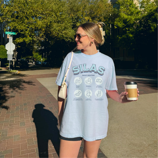 Woman wearing a graphic 'SILAS Daily Routine' tee with cat illustrations, holding coffee outdoors.