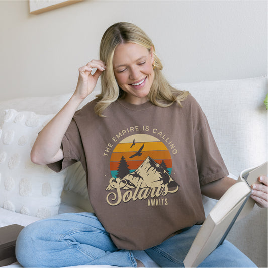 Woman reading on sofa wearing brown Solaris Awaits graphic tee with mountain sunset design