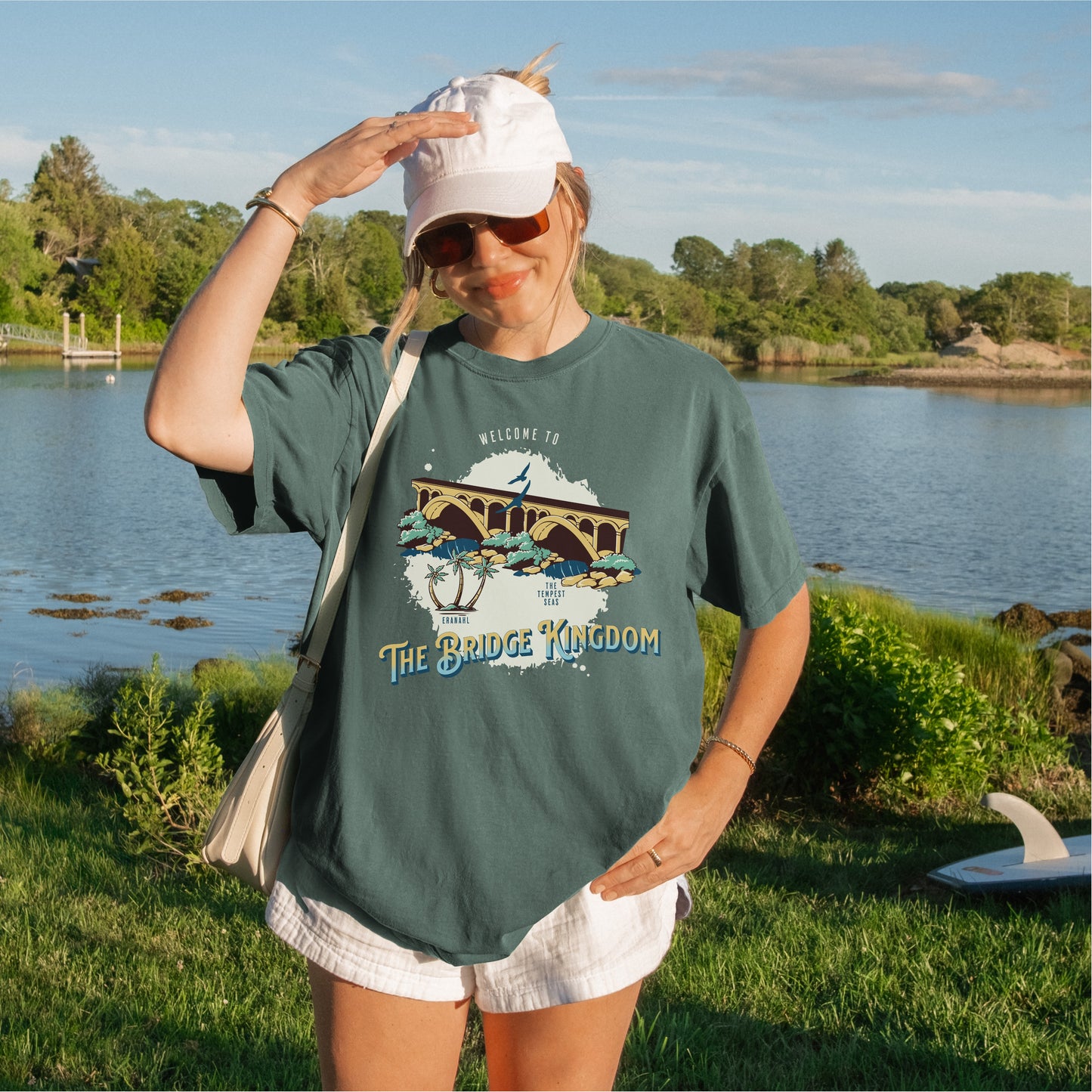 Woman outdoors by a lake wearing a green The Bridge Kingdom graphic T-shirt and white cap