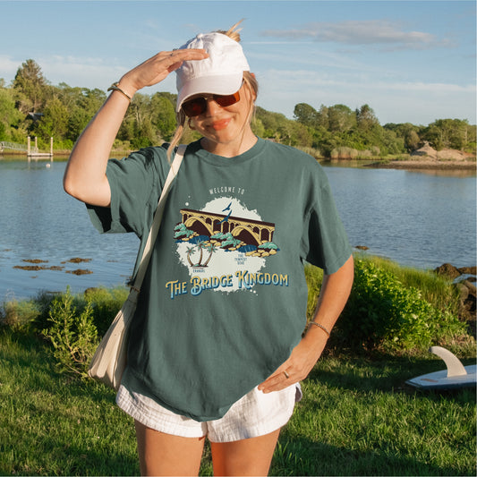 Woman outdoors by a lake wearing a green The Bridge Kingdom graphic T-shirt and white cap