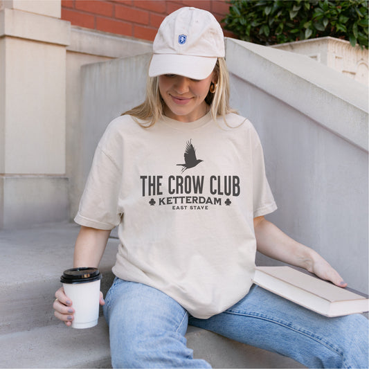 Woman wearing The Crow Club Ketterdam bookish tee, holding book and coffee cup on outdoor steps