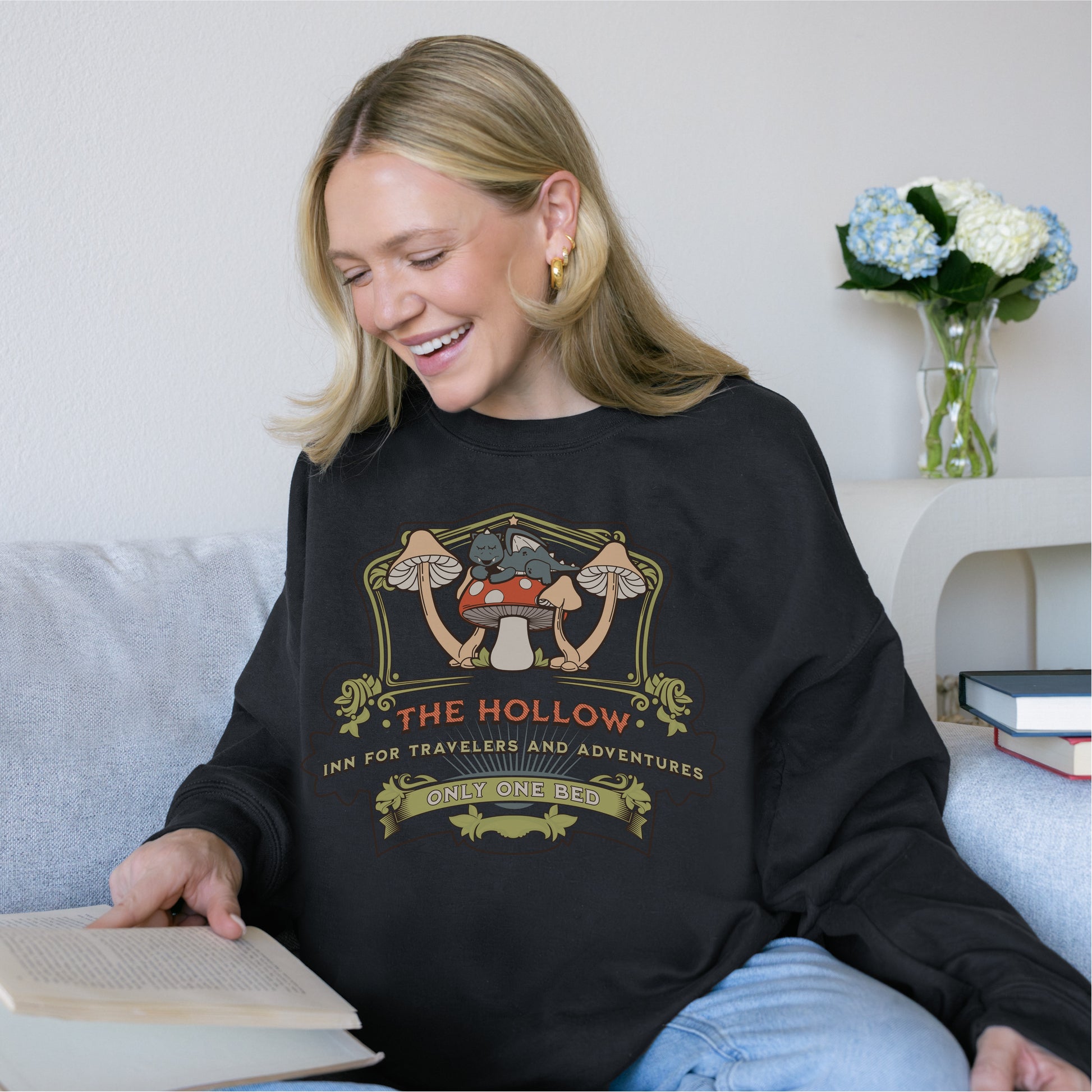 Smiling woman reading on couch in black bookish sweater with mushroom and cat graphic