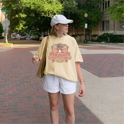 Woman wearing beige 'The Hollow' graphic bookish T-shirt, white shorts, and white cap outdoors
