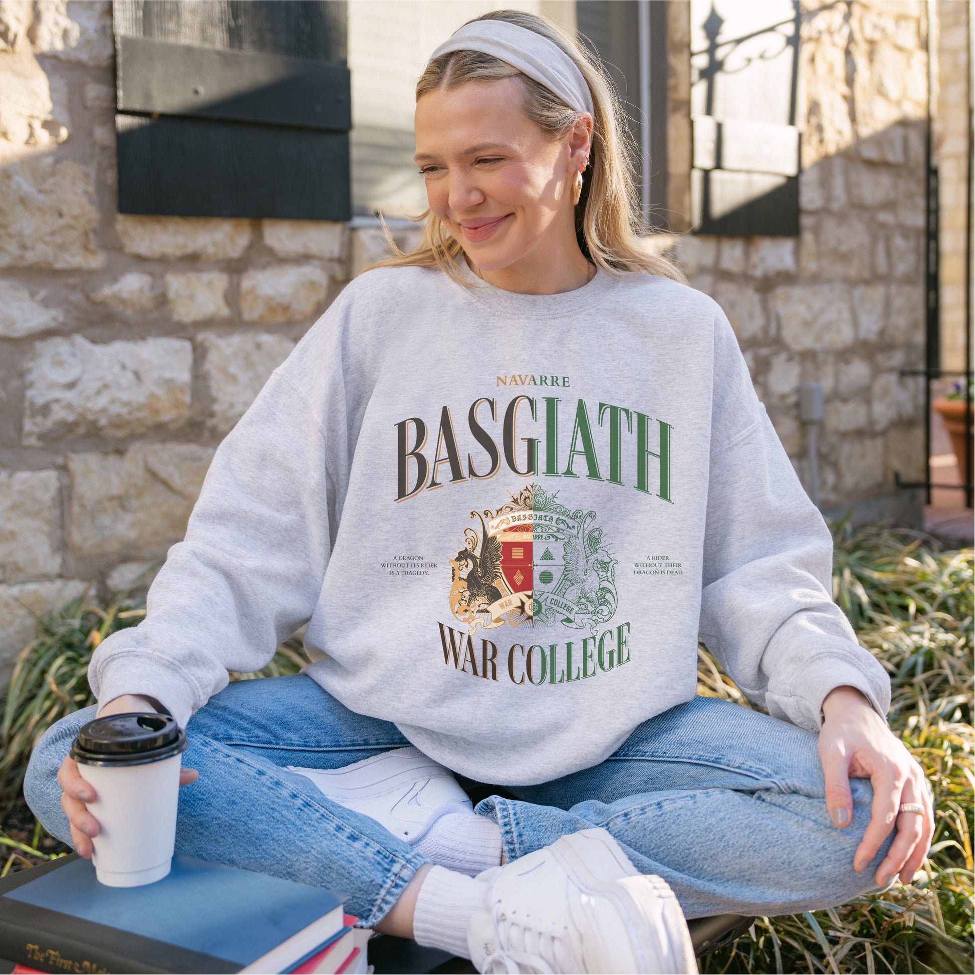 Woman in Basgiath War College sweatshirt sitting outside, holding coffee, wearing jeans and sneakers.