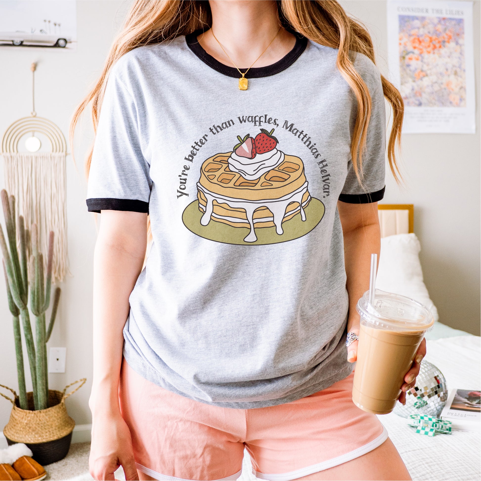 Woman wearing grey ringer tee with waffles graphic and quote, holding iced coffee in bedroom