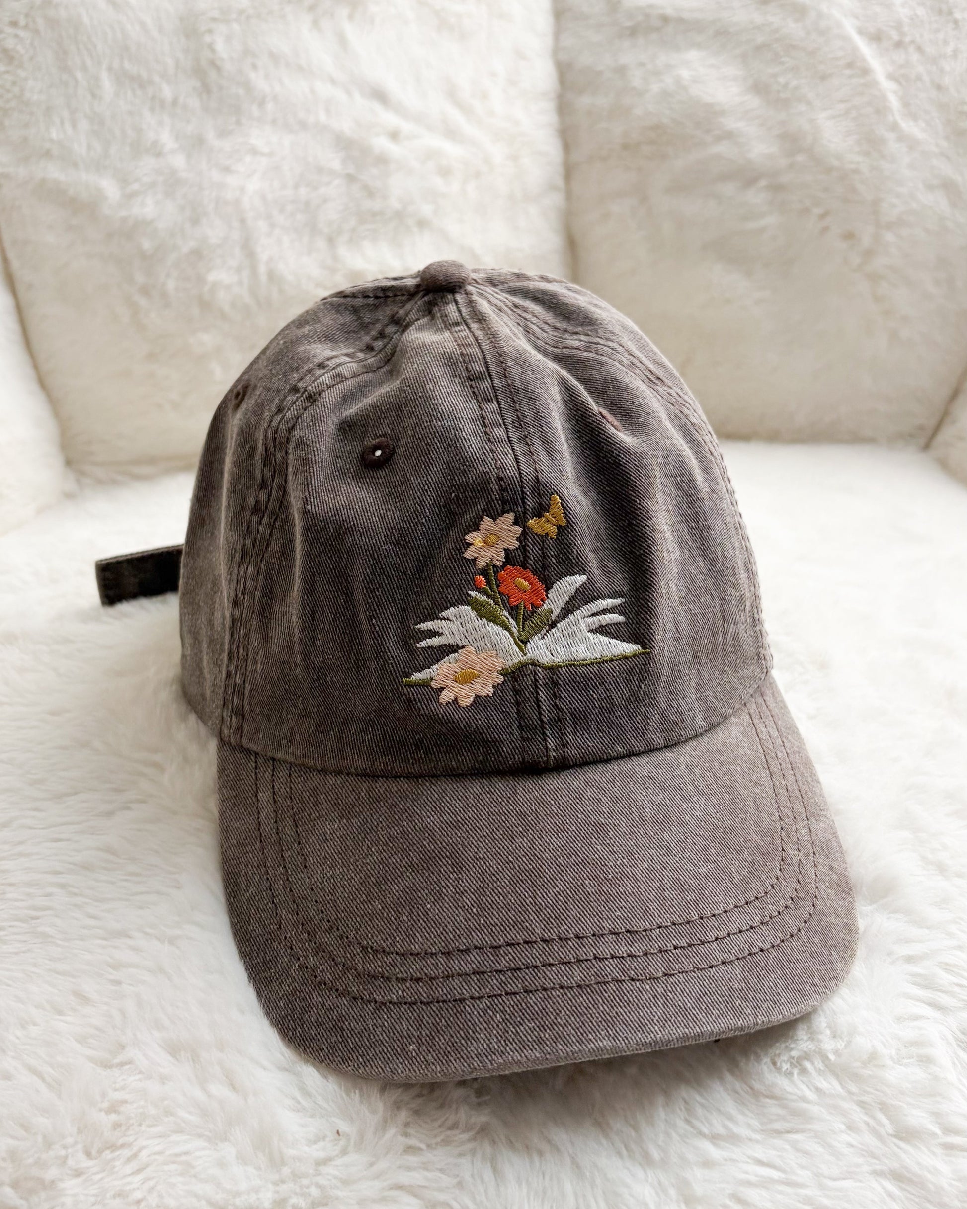 Brown embroidered baseball cap with flowers, butterfly, and book design on soft white background