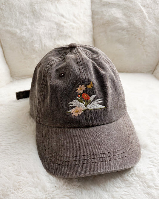 Brown embroidered baseball cap with flowers, butterfly, and book design on soft white background