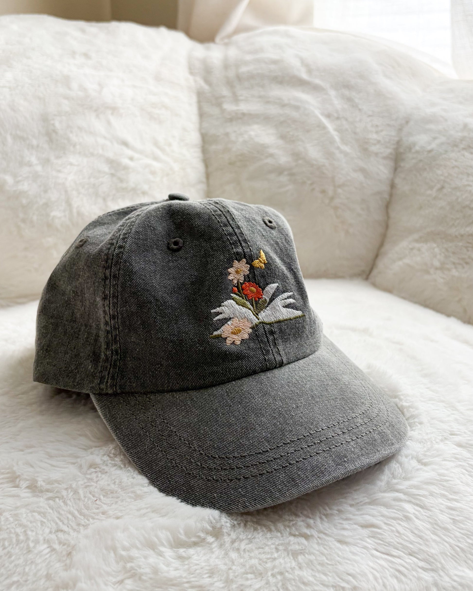 Grey denim baseball cap with embroidered hand holding wildflowers, resting on a plush white surface.