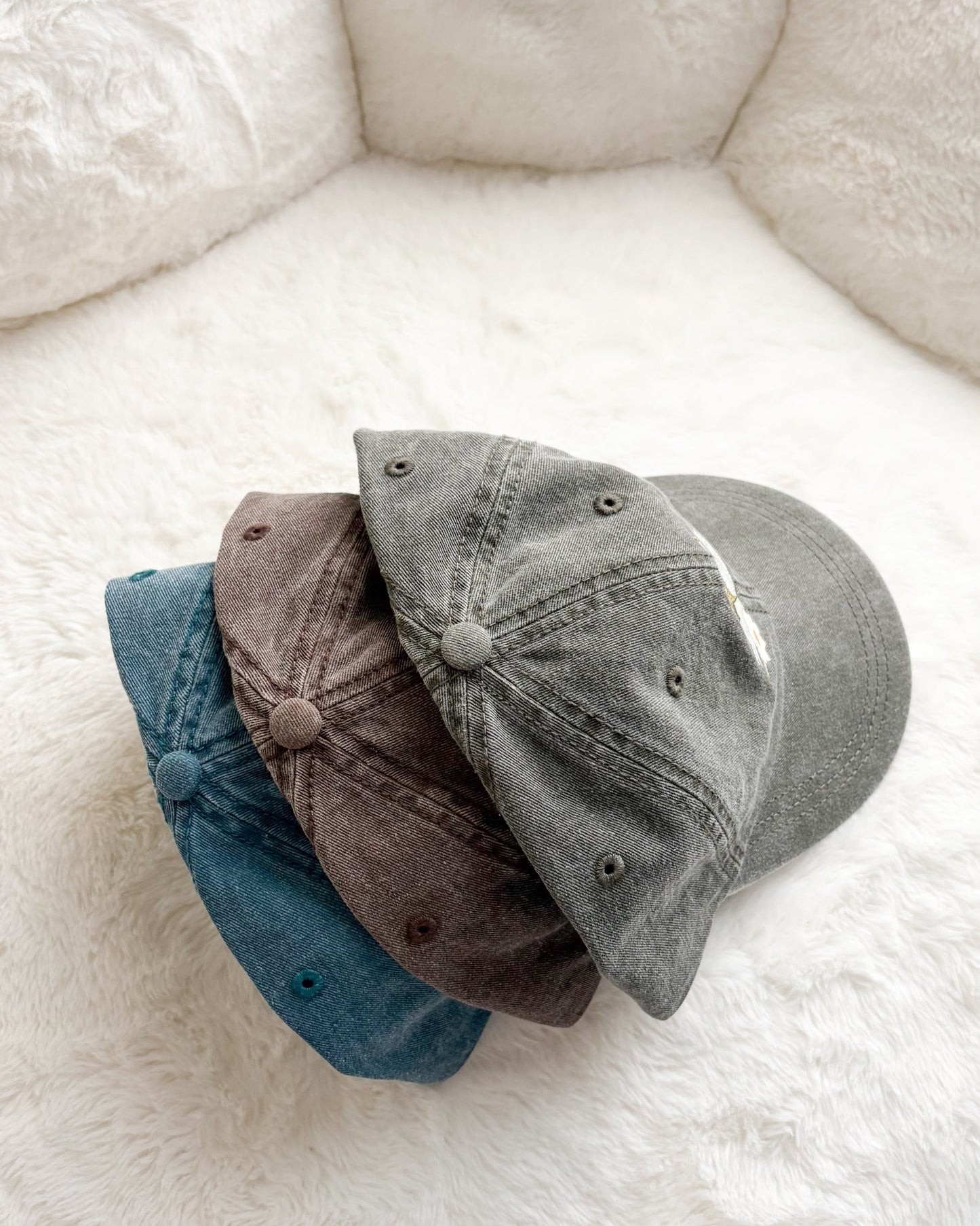 Three unstructured baseball caps in blue, brown, and grey on a fluffy white background