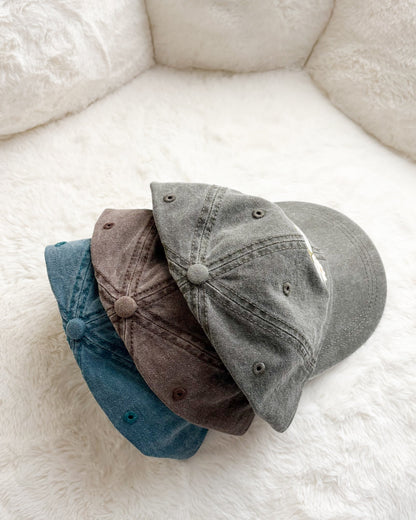 Three unstructured baseball caps in blue, brown, and grey on a fluffy white background