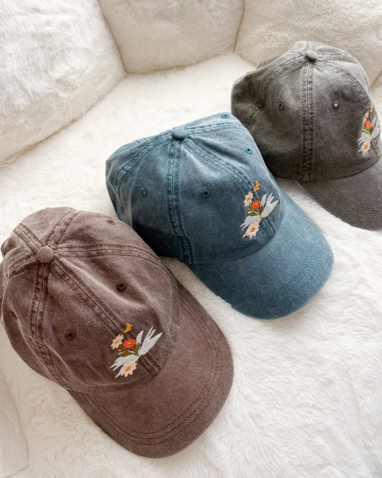 Three vintage-wash baseball caps with embroidered floral design on soft white blanket