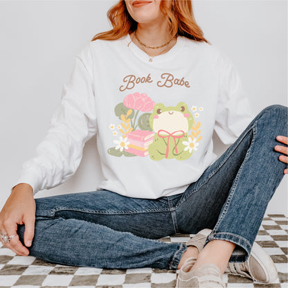Woman wearing white sweatshirt with 'Book Babe' text, cute frog, books, and flowers graphic.
