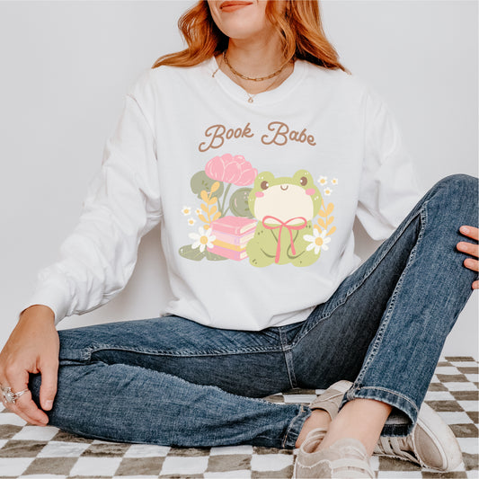 Woman wearing white sweatshirt with 'Book Babe' text, cute frog, books, and flowers graphic.