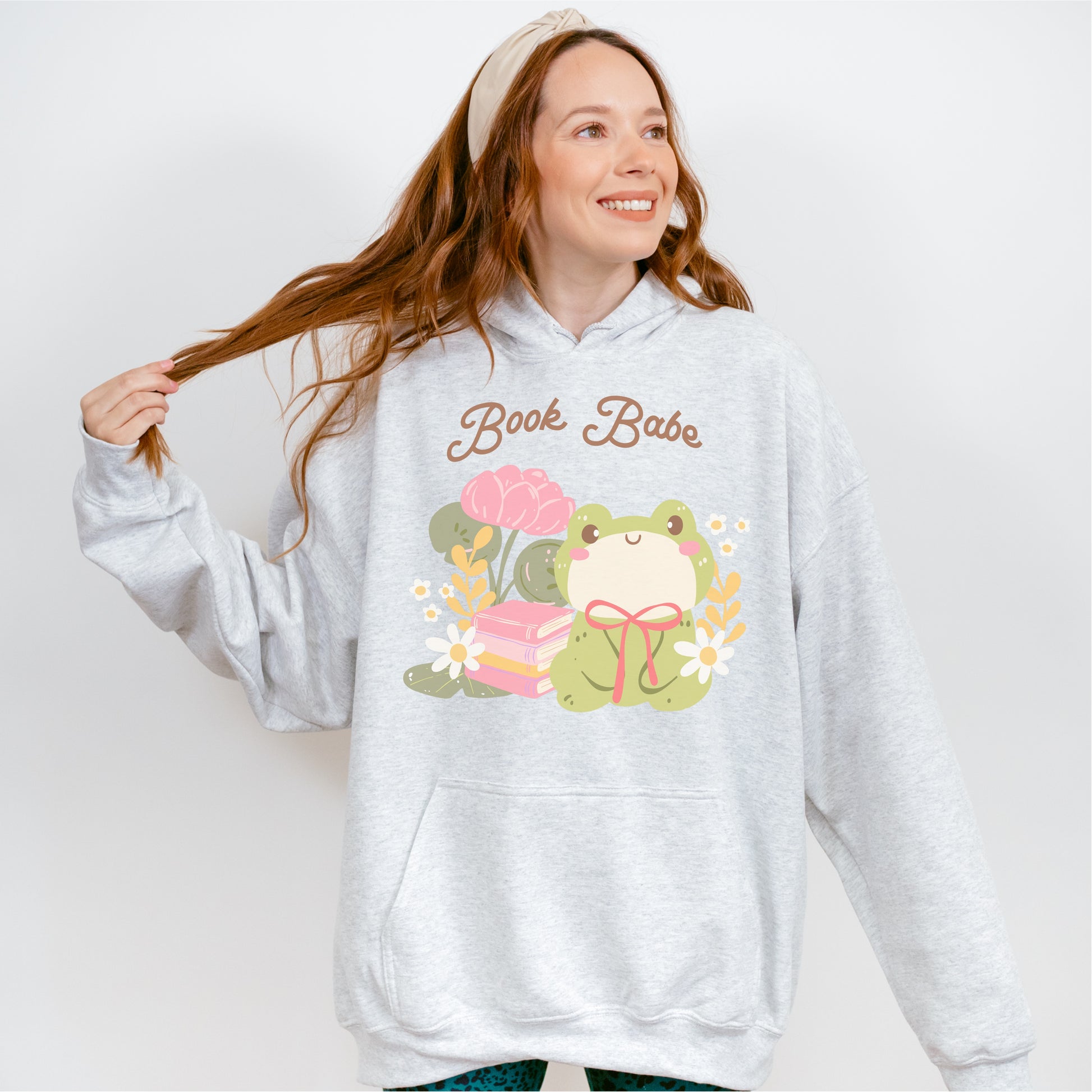 Woman wearing a gray hoodie with 'Book Babe' text and cute frog, books, and flowers graphic