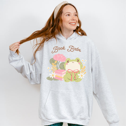 Woman wearing a gray hoodie with 'Book Babe' text and cute frog, books, and flowers graphic