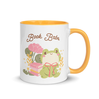 White ceramic mug with yellow handle, featuring a cute frog, flowers, books, and 'Book Babe' text.
