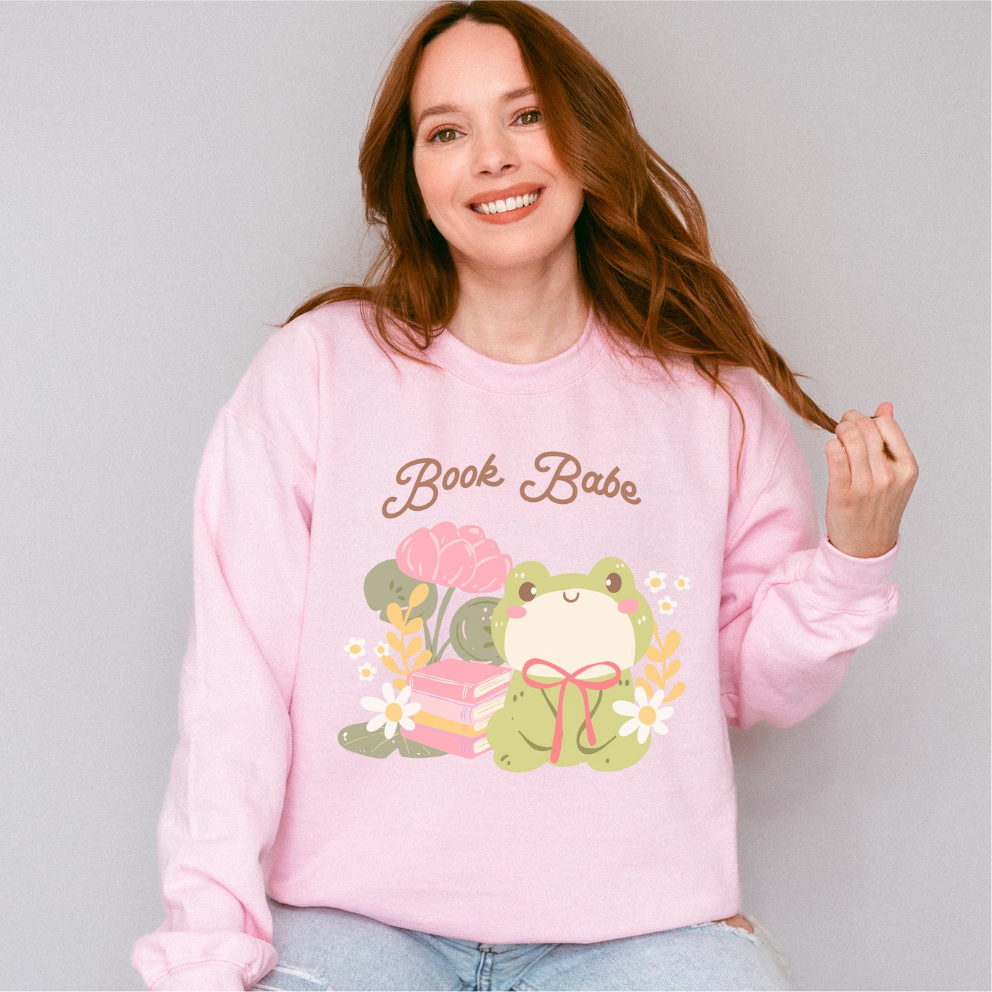 Woman wearing pink sweatshirt with 'Book Babe' text, cute frog, books, and flowers graphic