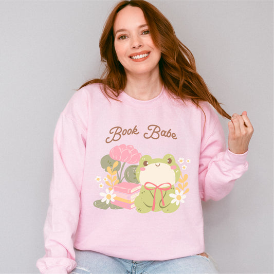 Woman wearing pink sweatshirt with 'Book Babe' text, cute frog, books, and flowers graphic
