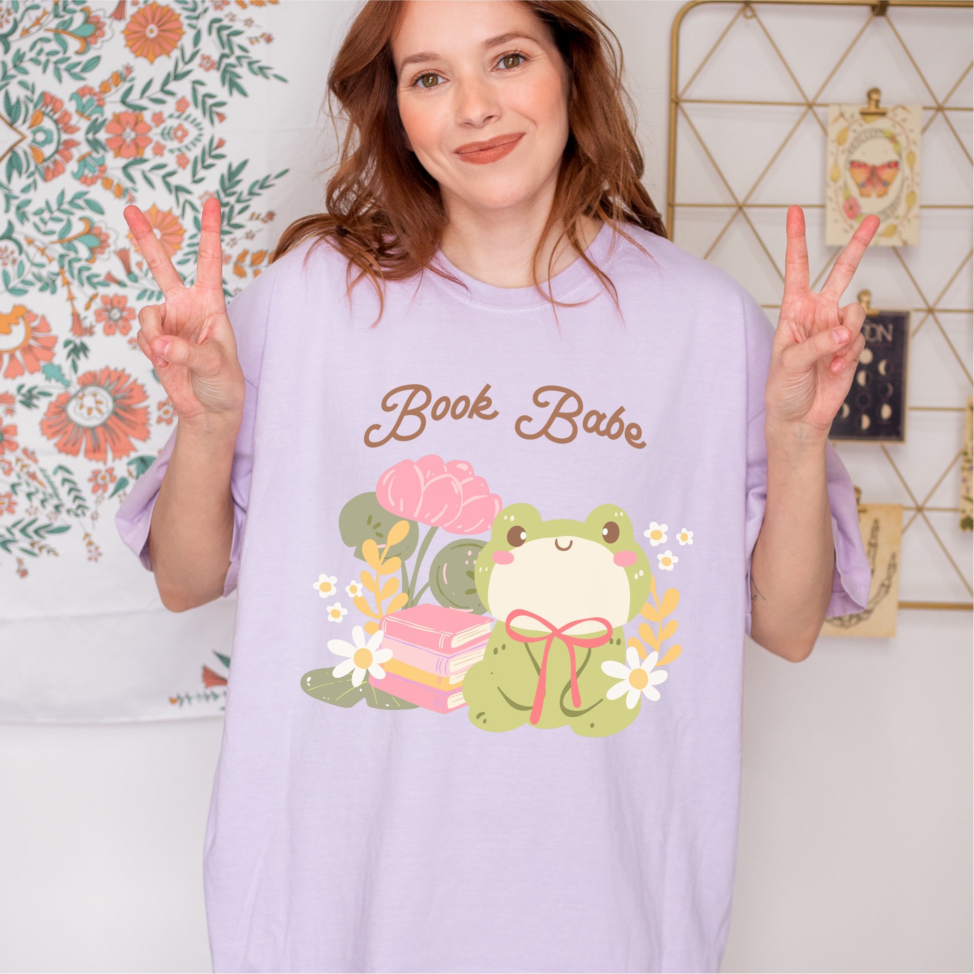 Woman wearing lilac Book Babe t-shirt with cute frog, books, flowers, and peace sign