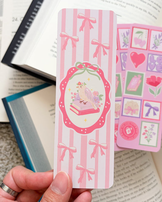 Hand holding a pink decorative bookmark with a design of a book and heart, against a background of open books.
