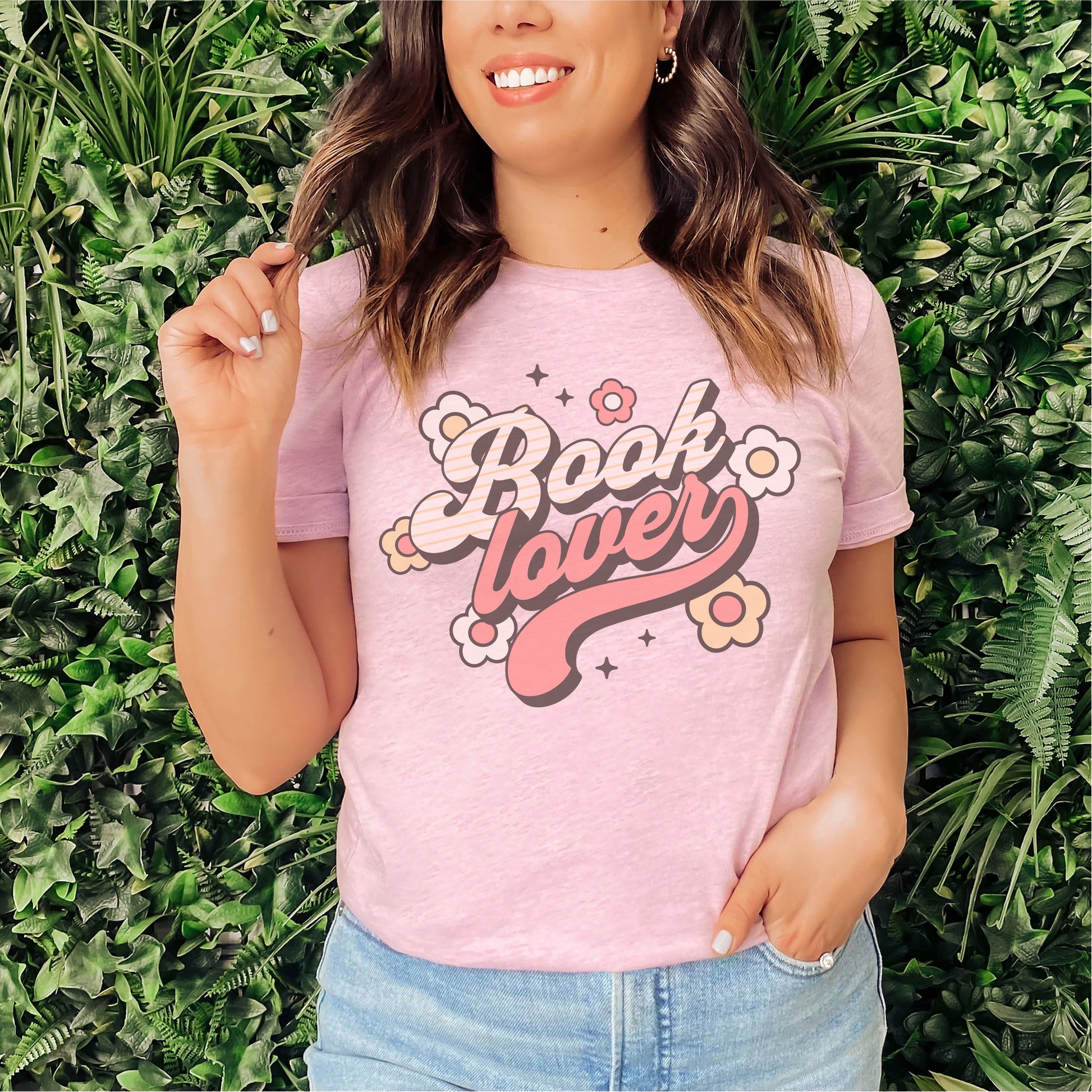 Woman wearing pink 'Book lover' graphic t-shirt with retro flower design, standing by green plants