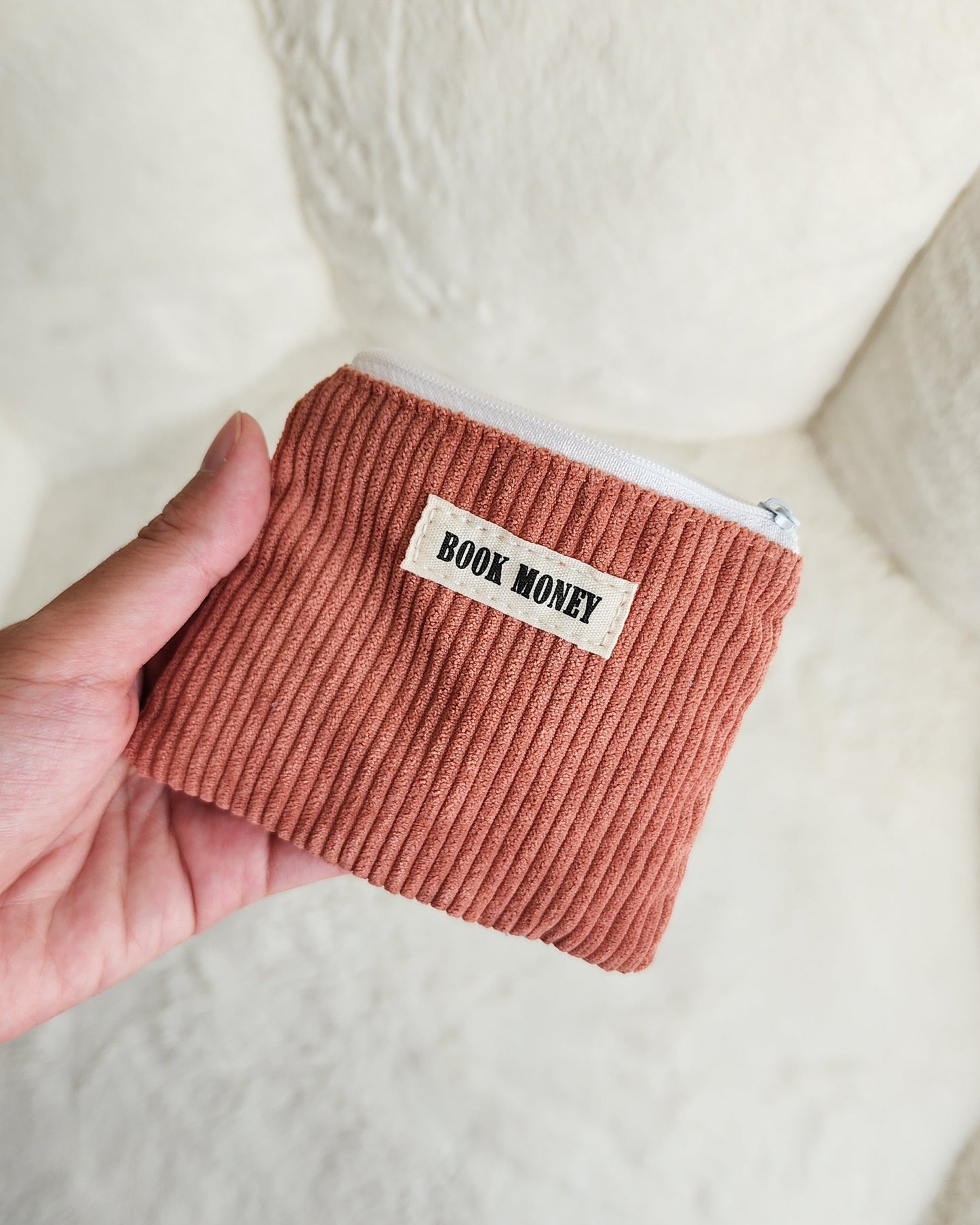 Hand holding a brown corduroy zippered coin purse with 'Book Money' label