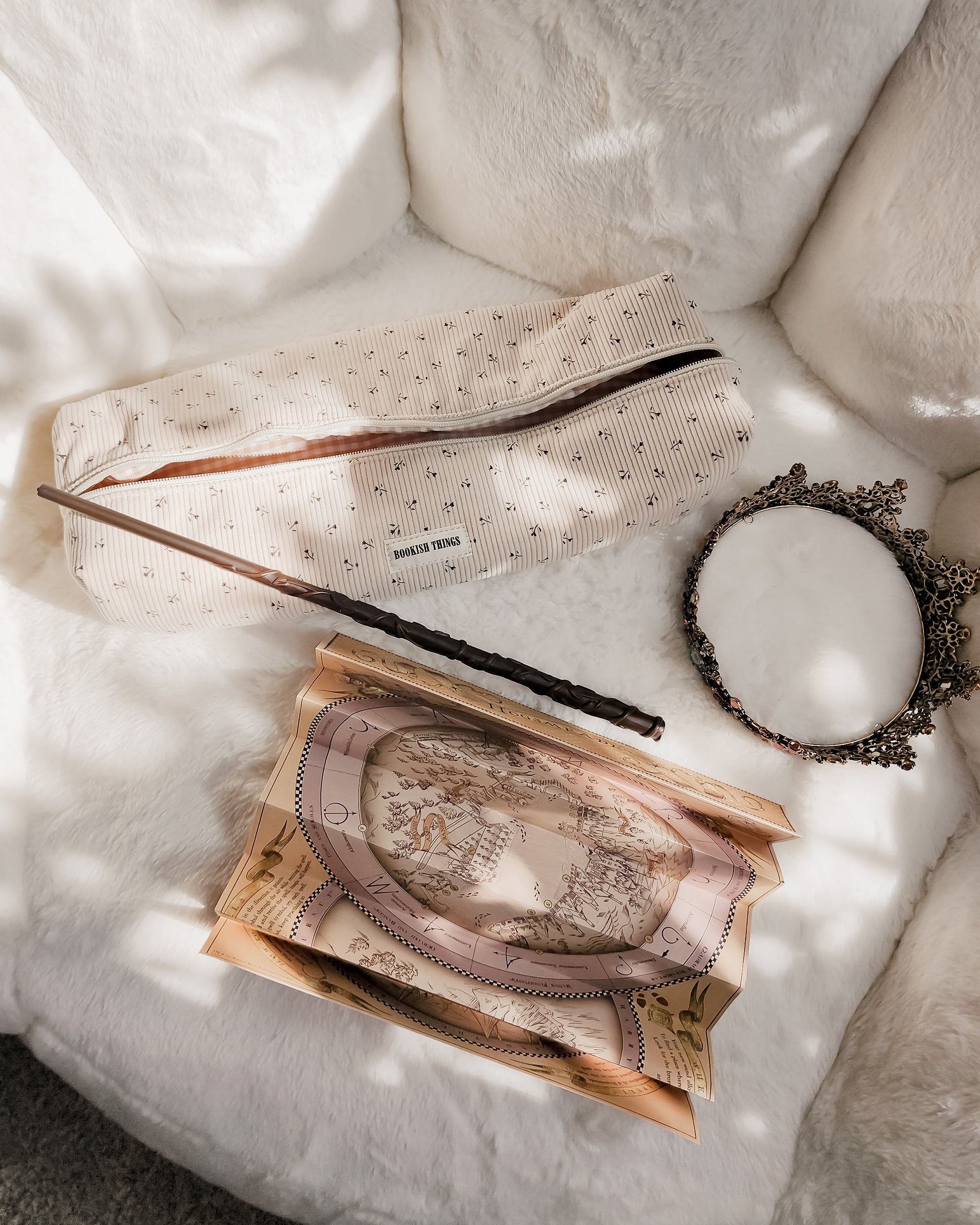 Beige corduroy travel bag labeled 'BOOKISH THINGS' with a wand, vintage map, and ornate mirror on a white plush surface.