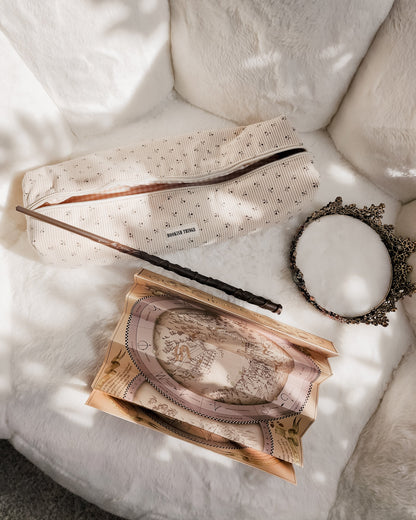 Beige corduroy travel bag labeled 'BOOKISH THINGS' with a wand, vintage map, and ornate mirror on a white plush surface.
