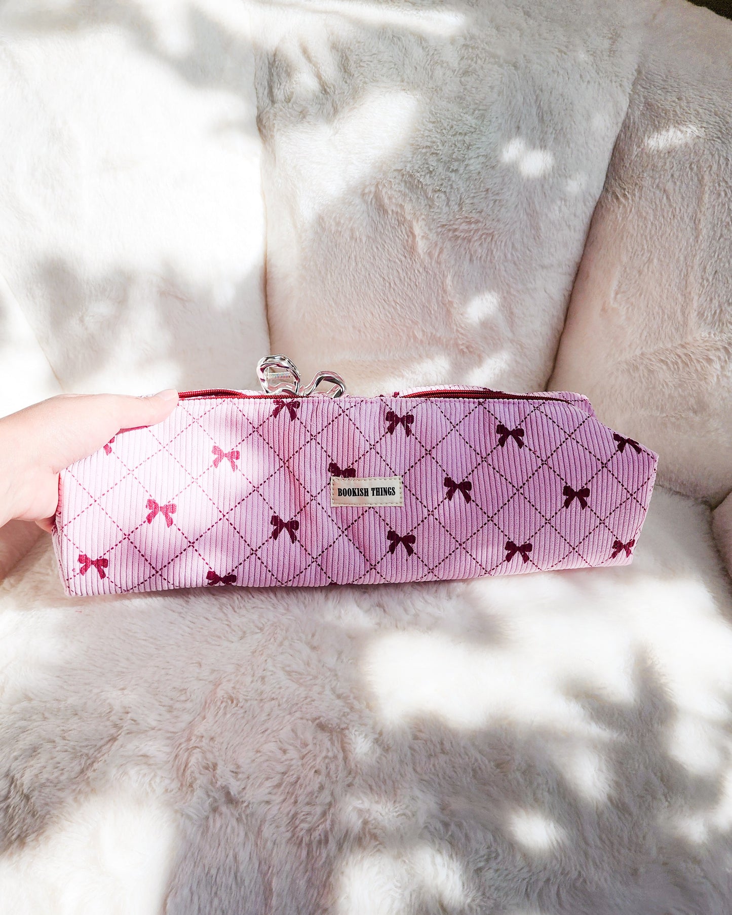 Pink corduroy travel bag with red bow patterns and 'Bookish Things' label on soft fabric chair