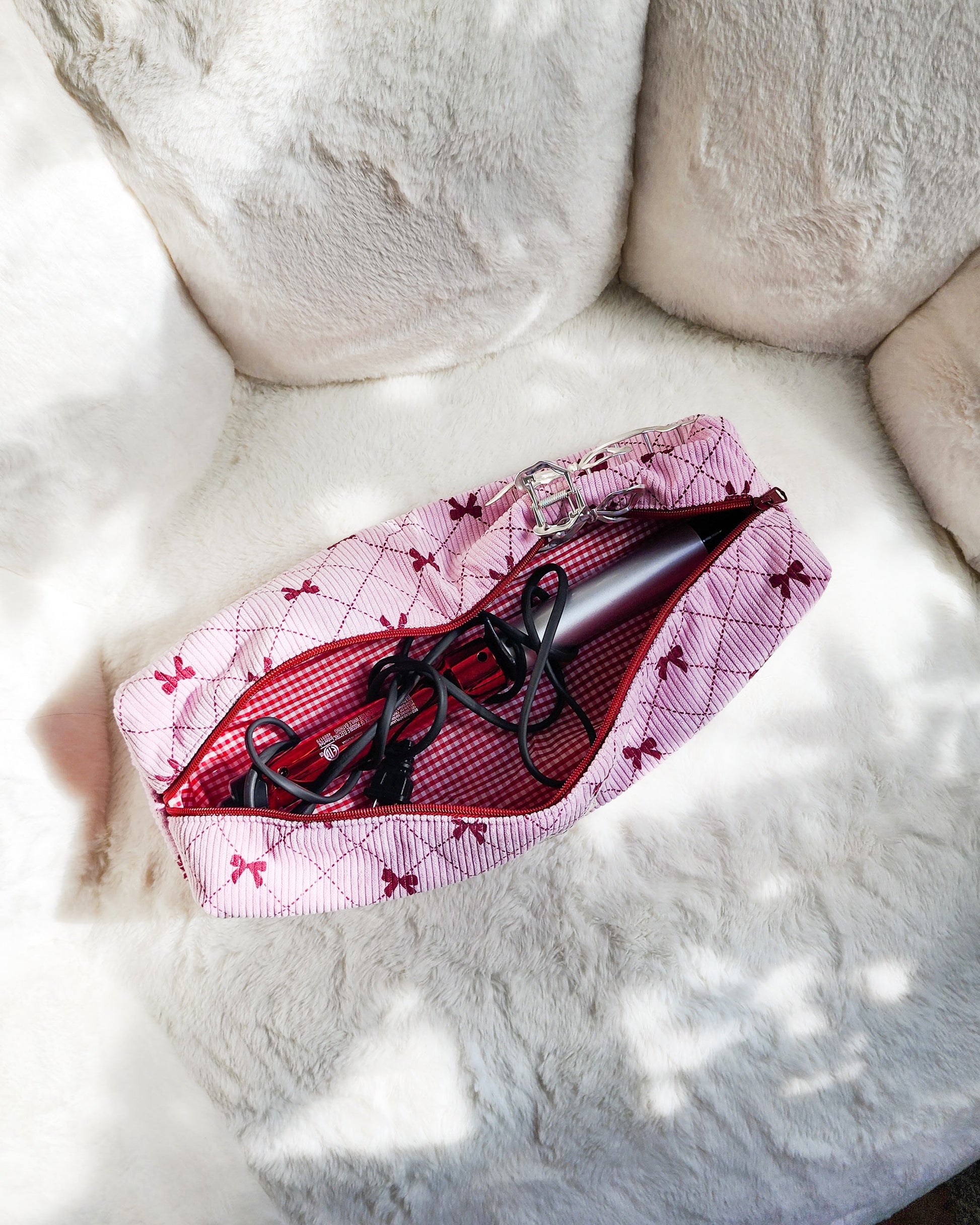 Pink corduroy hair tool travel bag with red bows holding a curling iron on a white plush surface