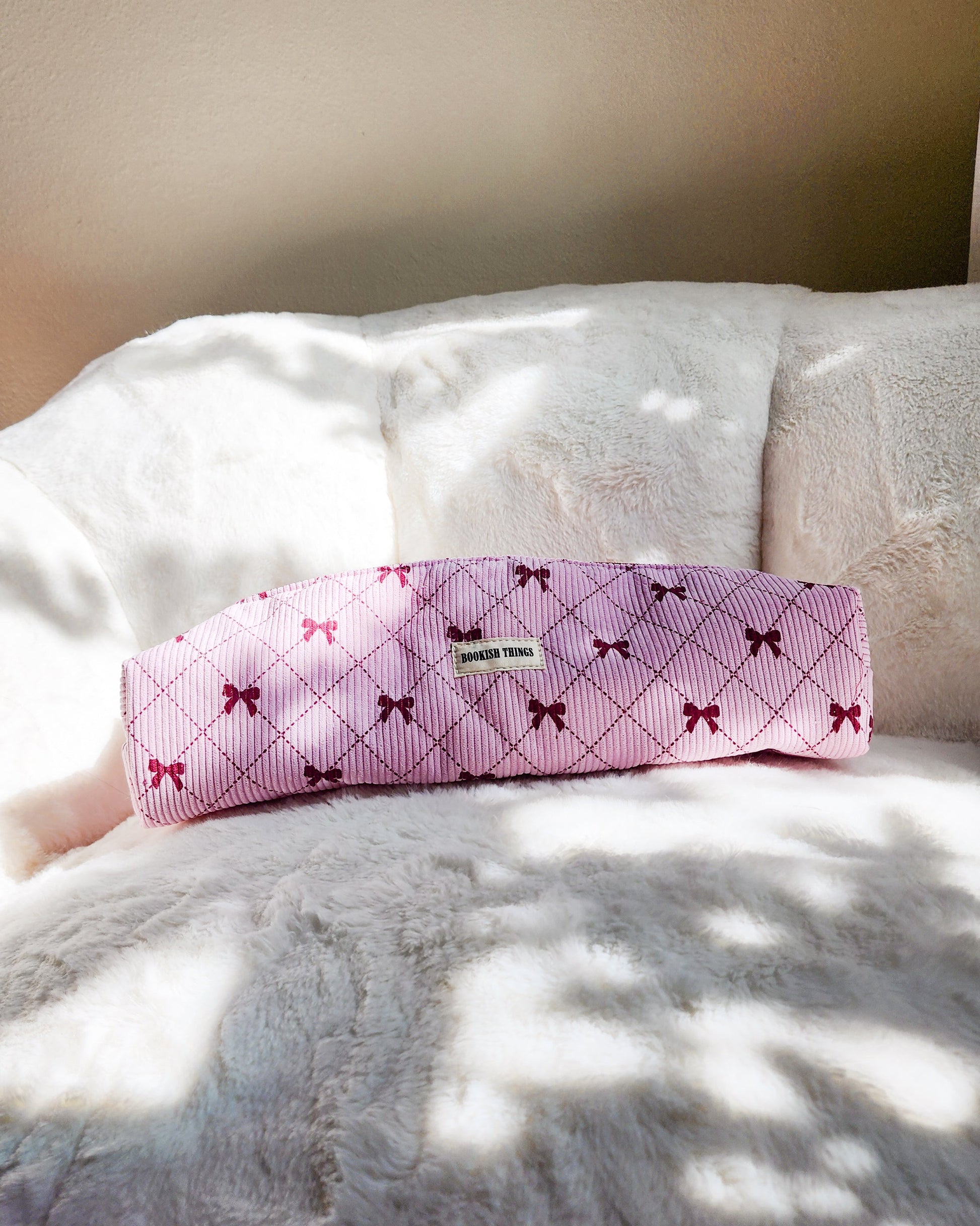 Pink corduroy travel bag with red bows and 'Bookish Things' label on a white plush blanket