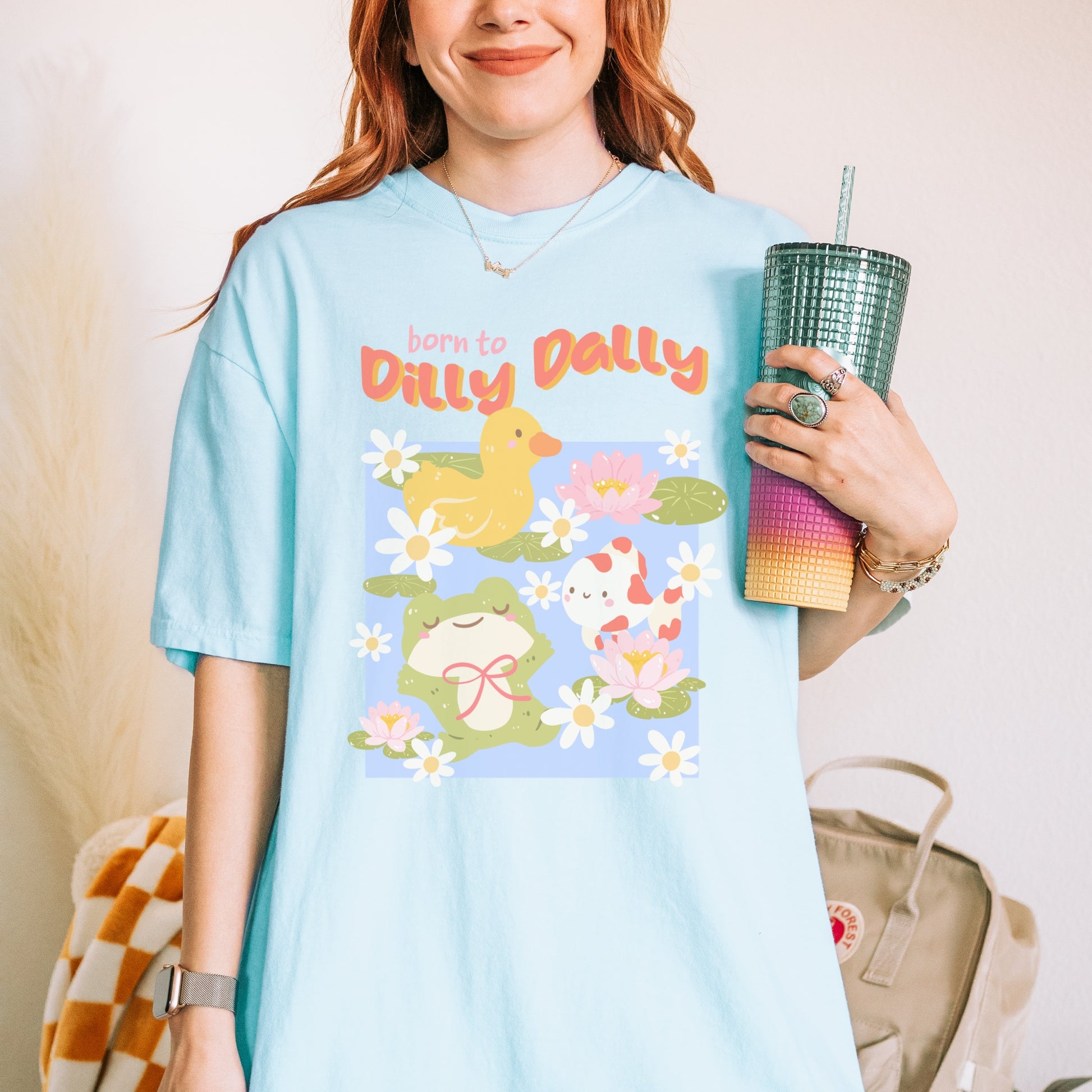 Light blue T-shirt with 'Born to Dilly Dally' text, featuring cute cartoon frog, duck, and koi pond design.
