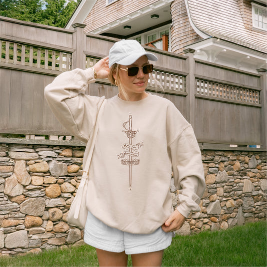 Woman wearing a beige bookish sweatshirt with sword and banner graphic, outdoors