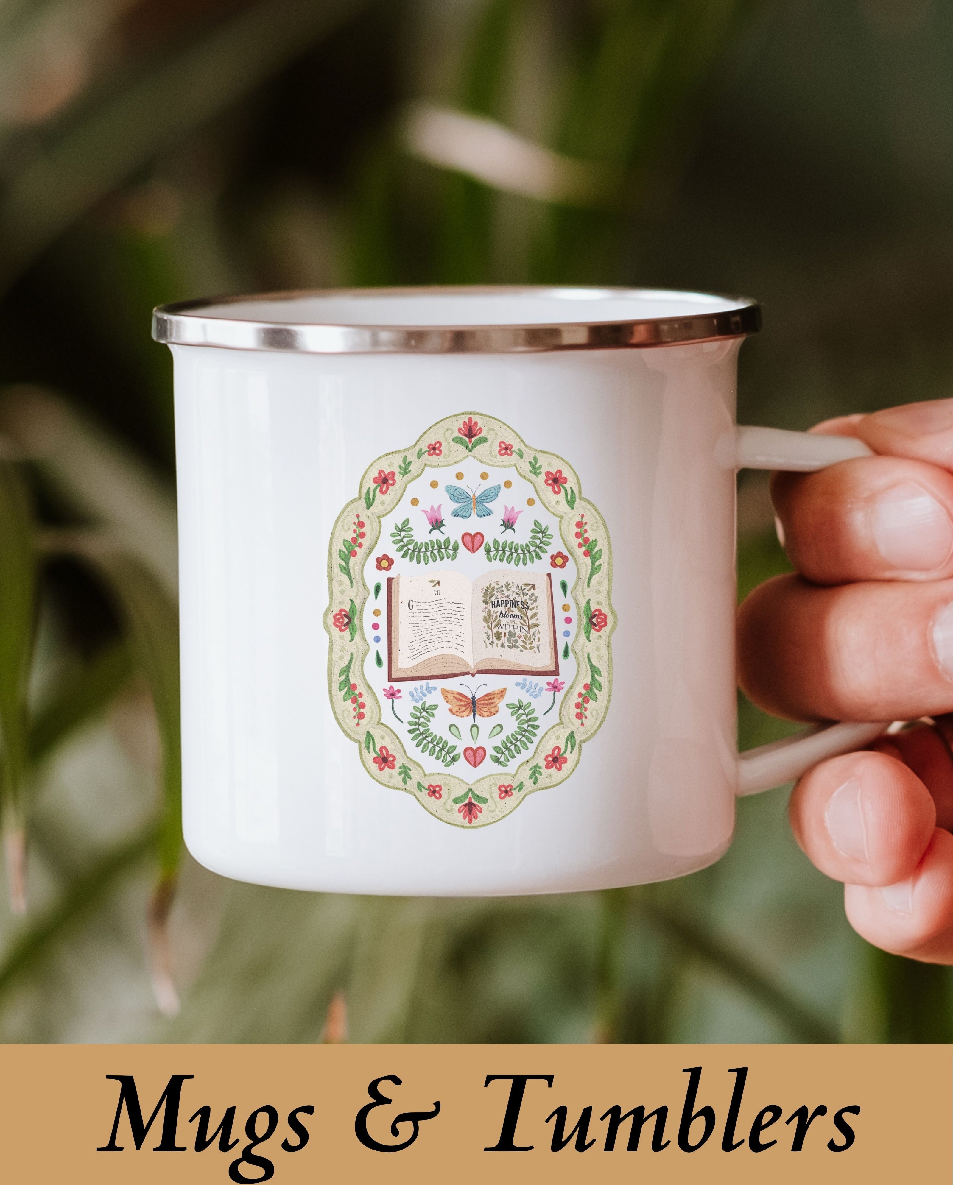 A hand holding a cozy, wholesome bookish enamel mug printed with a cottagecore floral and butterfly book illustration, styled outdoors against soft green foliage for a mindful reading aesthetic.