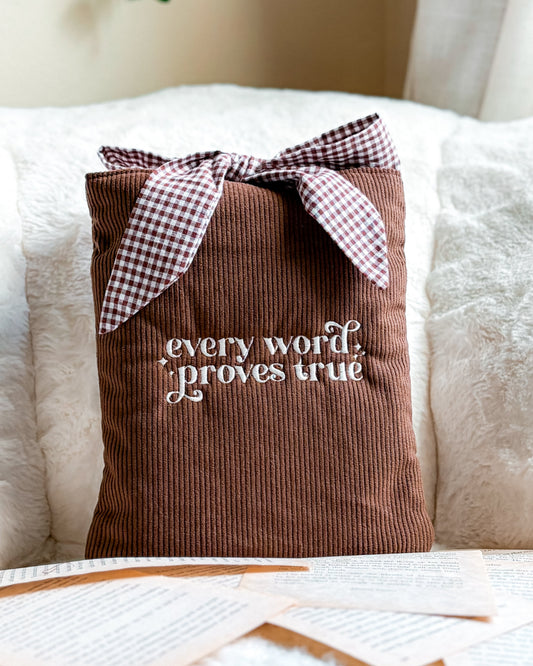 Brown corduroy book sleeve with embroidered quote and checkered bow, surrounded by book pages
