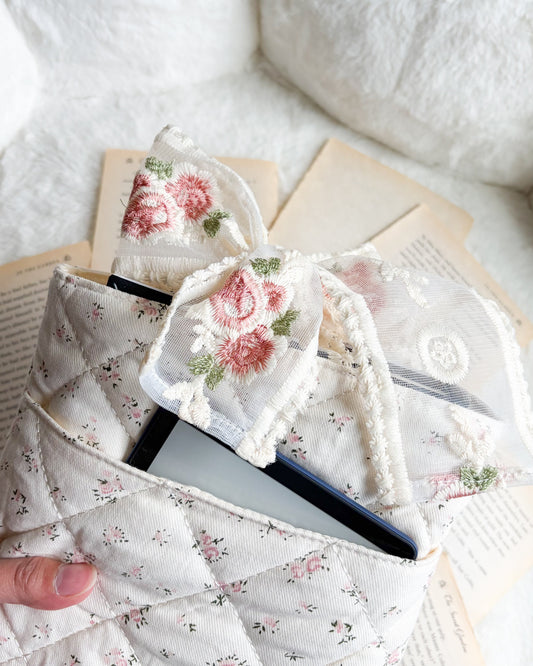 Floral quilted pouch with embroidered bow holding an e-reader, surrounded by book pages.