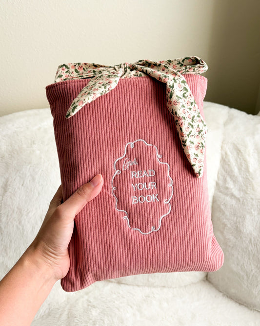 Pink book sleeve with 'Read Your Book' embroidery and floral ribbon held by a hand on a white surface.