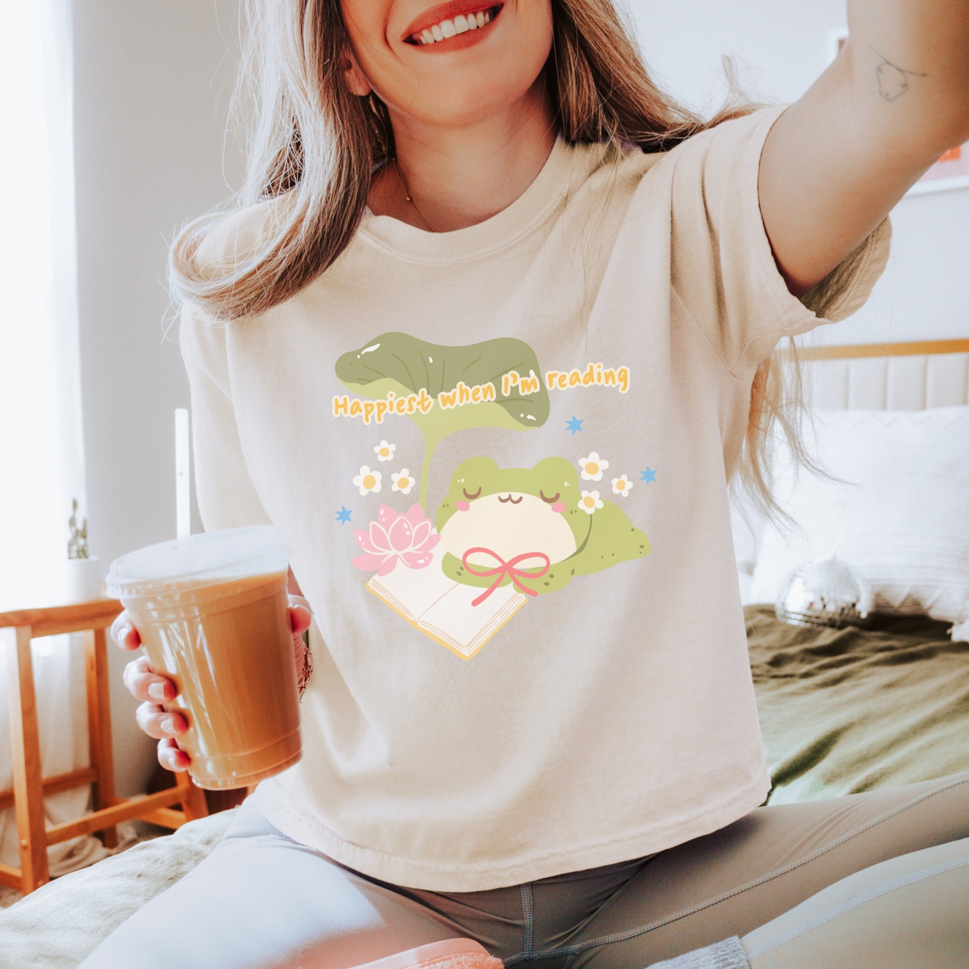 Woman wearing a cute frog reading shirt with 'Happiest when I'm reading' text, holding iced coffee